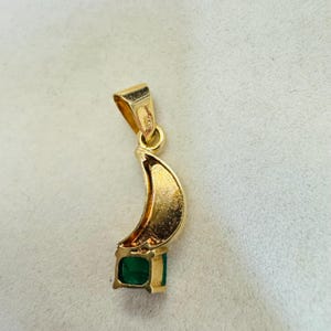 K&ouml;nnte beinhalten: Goldener Anh&auml;nger mit einem Halbmond-Design und einem quadratischen Smaragd-Edelstein. Der Anh&auml;nger hat oben eine Schlaufe f&uuml;r eine Kette. Der Smaragd hat eine tiefgr&uuml;ne Farbe.