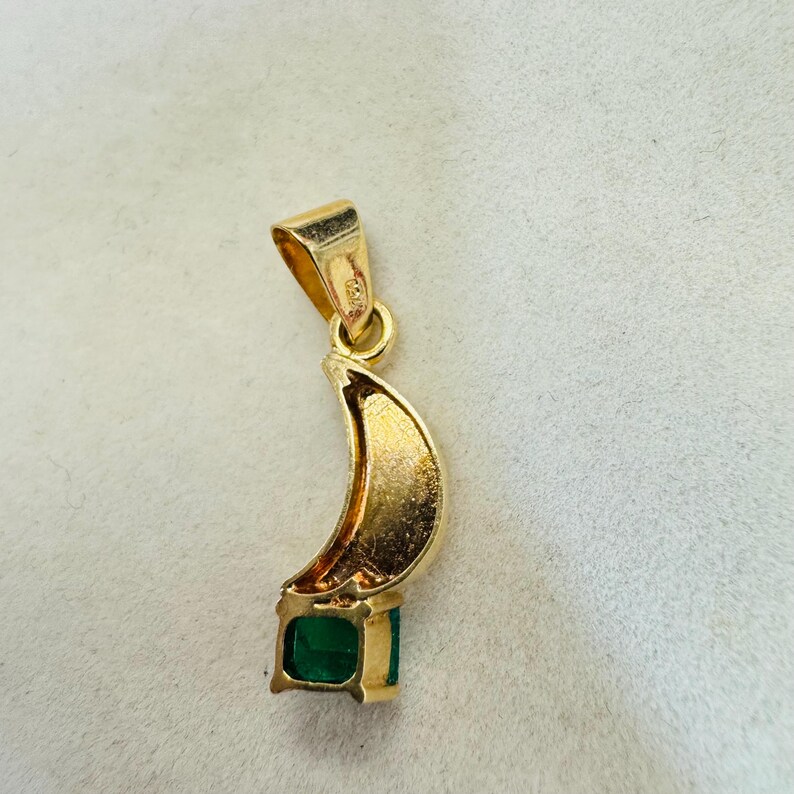 K&ouml;nnte beinhalten: Goldener Anh&auml;nger mit einem Halbmond-Design und einem quadratischen Smaragd-Edelstein. Der Anh&auml;nger hat oben eine Schlaufe f&uuml;r eine Kette. Der Smaragd hat eine tiefgr&uuml;ne Farbe.