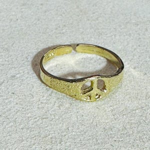 Puede incluir: Un anillo de tono dorado con un diseño de banda abierta y superficie texturizada. El punto focal del anillo es un símbolo de paz recortado integrado en la banda, que muestra un acabado metálico y una textura martillada ligeramente rugosa.