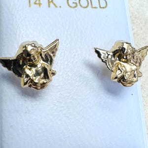 Op de afbeelding: Een paar goudkleurige engel oorknopjes. Elke oorbel toont een engel met vleugels. De oorbellen worden gepresenteerd op een witte achtergrond, met de tekst "14 K. GOLD" zichtbaar.