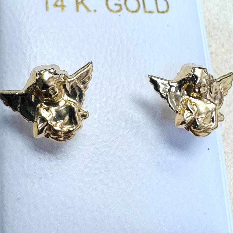 Op de afbeelding: Een paar goudkleurige engel oorknopjes. Elke oorbel toont een engel met vleugels. De oorbellen worden gepresenteerd op een witte achtergrond, met de tekst "14 K. GOLD" zichtbaar.