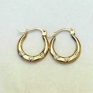 Könnte beinhalten: Ein Paar polierte Gold-Creolen mit einer Reihe kleiner, eingravierter Herzformen auf der Außenseite jedes Reifens. Die Ohrringe sind mit einem Klappverschluss ausgestattet, der einen sicheren Halt der kreisförmigen Creolen gewährleistet.