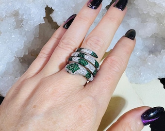 May include: A silver snake ring with white and green gemstones. The ring coils around the finger, with the snake's head and tail detailed with green stones. The body of the snake is adorned with white gemstones.