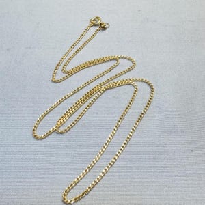 Op de afbeelding: Een goudkleurige metalen ketting bestaande uit een reeks kleine, in elkaar grijpende ovale schakels. De ketting is dun en fijn, met aan het uiteinde een rond veerringslotje dat is ontworpen om de ketting veilig om de nek te sluiten.