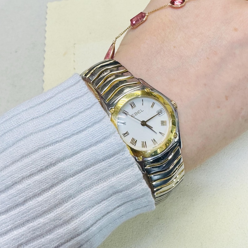 Pode incluir: Um rel&oacute;gio Ebel bicolor com mostrador branco, moldura dourada e pulseira prateada e dourada. O rel&oacute;gio apresenta algarismos romanos e um visor de data. Uma pulseira de ouro com pedras preciosas rosa tamb&eacute;m &eacute; vis&iacute;vel.