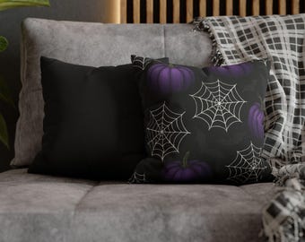 May include: Two decorative throw pillows on a gray couch. One pillow is solid black, and the other features a black background with white spiderwebs and purple pumpkins. A plaid blanket is draped over the back of the couch.