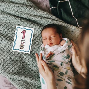 Peut inclure: Un nouveau-n&eacute; envelopp&eacute; dans une couverture blanche &agrave; motifs de feuilles vertes, doucement tenu. Une carte avec le chiffre "1", le mot "semaine" et un lapin de dessin anim&eacute; est plac&eacute;e &agrave; proximit&eacute;. Le b&eacute;b&eacute; dort paisiblement.