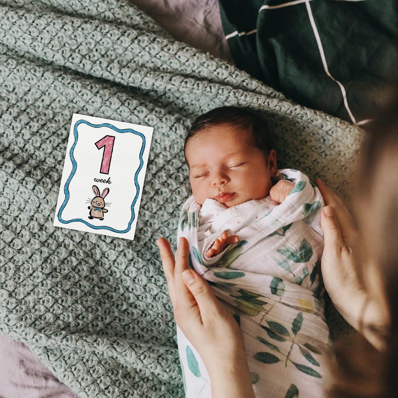 Peut inclure: Un nouveau-n&eacute; envelopp&eacute; dans une couverture blanche &agrave; motifs de feuilles vertes, doucement tenu. Une carte avec le chiffre "1", le mot "semaine" et un lapin de dessin anim&eacute; est plac&eacute;e &agrave; proximit&eacute;. Le b&eacute;b&eacute; dort paisiblement.