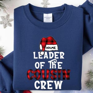 May include: Navy blue sweatshirt with the text "LEADER OF THE COUSIN CREW" in white and red plaid lettering. A Santa hat graphic with the word "NAME" is above the text. The sweatshirt is a crew neck.