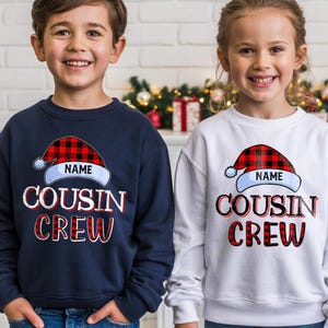 May include: Two children wearing navy blue and white sweatshirts. Each sweatshirt features a Santa hat design with the word "NAME" above "COUSIN CREW" in red and white lettering. The sweatshirts are likely for a holiday or family gathering.