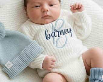 May include: A newborn baby wearing a white knit onesie with the name "Barney" embroidered in blue. The baby is also wearing a light blue knit hat with a pom-pom and matching booties. The baby is lying on a white blanket.