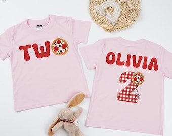 Puede incluir: Dos camisetas rosas de manga corta. Una camiseta tiene la palabra "TWO" y un gráfico de pizza. La otra camiseta tiene el nombre "OLIVIA" y el número "2" con un gráfico de pizza. Un conejito de peluche está en la imagen.