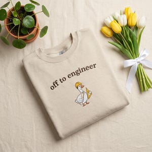 May include: Beige sweatshirt with the text "off to engineer" and an embroidered cartoon goose wearing a yellow hard hat and holding a triangle ruler. A potted plant and a bouquet of yellow and white tulips are in the background.