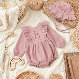 May include: A dusty rose baby romper with long sleeves and ruffle detailing, paired with a matching sun hat. The romper features a large letter "E" and the name "Evelyn" embroidered in a glittery script. The hat also has the name "Evelyn" embroidered on it.