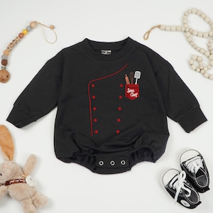 May include: Black baby romper with long sleeves, featuring a red chef coat design. The design includes a red pocket with cooking utensils and the text "Sous Chef". The romper has a button closure at the bottom.