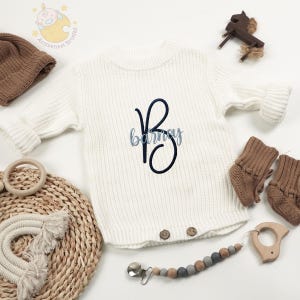 May include: A white knit baby romper with the name "Barney" embroidered in blue script. The romper has a round neck and button closures. Accessories include a brown knit hat, booties, and a wooden teether.