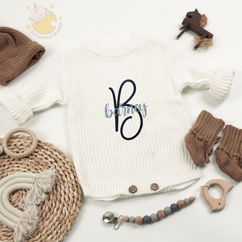 May include: A white knit baby romper with the name "Barney" embroidered in blue script. The romper has a round neck and button closures. Accessories include a brown knit hat, booties, and a wooden teether.
