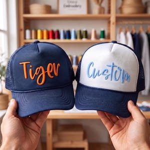 May include: Two trucker hats with mesh backs. The left hat is navy blue with the word Tiger in raised orange lettering. The right hat has a white front panel and navy blue brim with the word Custom in raised light blue lettering.