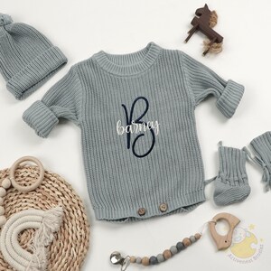 May include: A light blue knitted baby outfit set, including a sweater with the name "Barney" embroidered on it, a matching hat, booties, and a pacifier clip. The set is displayed with a wooden horse toy and a wooden teether.