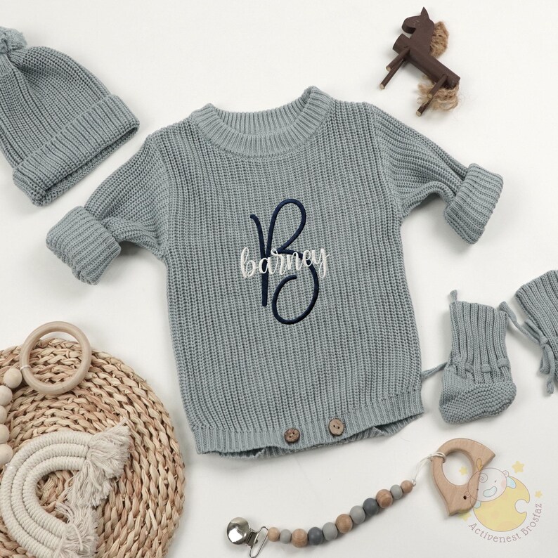 May include: A light blue knitted baby outfit set, including a sweater with the name "Barney" embroidered on it, a matching hat, booties, and a pacifier clip. The set is displayed with a wooden horse toy and a wooden teether.