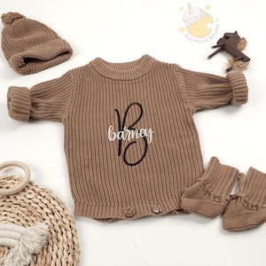 May include: A brown knit baby outfit set, including a sweater with the name "Barney" embroidered on the front, a matching beanie, and booties. The sweater has button details and ribbed texture.
