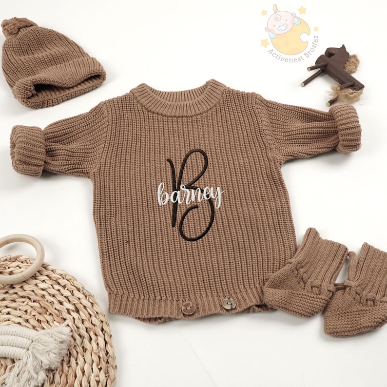 May include: A brown knit baby outfit set, including a sweater with the name "Barney" embroidered on the front, a matching beanie, and booties. The sweater has button details and ribbed texture.