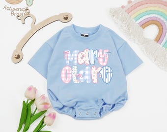 May include: A pale blue baby romper featuring the name "Mary Clare" in pink gingham, striped, and floral print lettering. The romper has short sleeves and popper fastenings. A wooden hanger and a rainbow decoration are also visible.