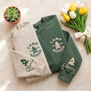 May include: Two sweatshirts, one beige and one green, with the text "CAMP BARKER" and a campfire graphic. The beige sweatshirt has "Charlie" embroidered on the sleeve. A small potted succulent and a bouquet of yellow and white tulips are also in the image.