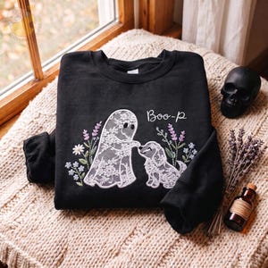 May include: A black crewneck sweatshirt featuring an embroidered design of a white lace ghost and a white lace dog surrounded by colorful floral sprigs. The text "Boo-p" is embroidered in white script above the ghost and dog figures.