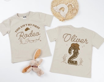 May include: Two beige t-shirts with cowboy-themed designs. One shirt reads "This ain't my first Rodeo, It's my 2nd" with a cowboy hat graphic. The other shirt has the name "Oliver" and a number 2 with a cowboy hat. A plush bunny is in the foreground.