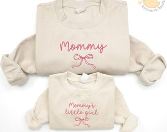 May include: Two beige sweatshirts, one adult and one child size, with pink embroidered text and bow designs. The adult sweatshirt says "Mommy," and the child's says "Mommy's little girl." A matching set for mother and child.