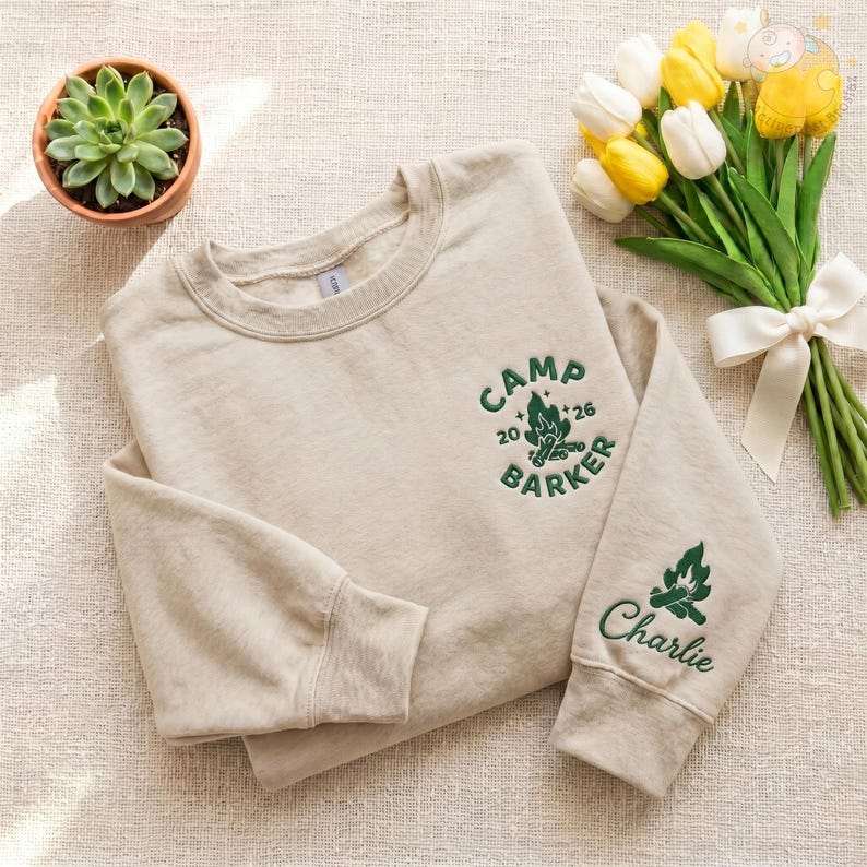 May include: Beige sweatshirt with green embroidered text and graphics. The text on the chest reads "CAMP BARKER 26". The sleeve features a campfire graphic and the name "Charlie". A small potted succulent and a bouquet of tulips are also in the image.