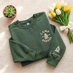 May include: A dark green sweatshirt with the text "CAMP BARKER 26" and a campfire graphic. The sleeve features a campfire graphic and the name "Charlie". A small potted succulent and a bouquet of yellow and white tulips are in the background.