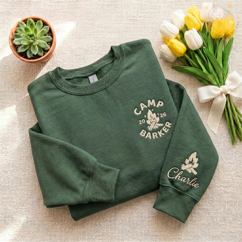 May include: A dark green sweatshirt with the text "CAMP BARKER 26" and a campfire graphic. The sleeve features a campfire graphic and the name "Charlie". A small potted succulent and a bouquet of yellow and white tulips are in the background.