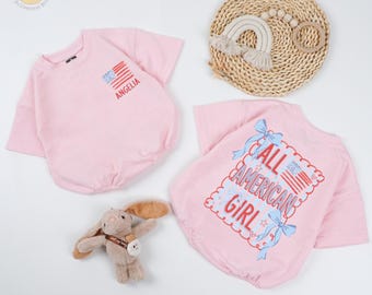 May include: Two pink baby rompers with short sleeves. One has an American flag graphic and the name "ANGELIA". The other says "ALL AMERICAN GIRL" with an American flag graphic. A stuffed bunny and a rainbow-shaped toy are also in the image.