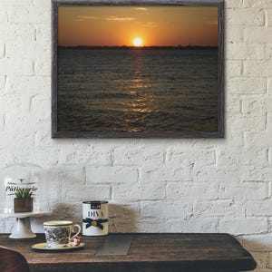 May include: A framed print depicting a sunset over water. The sun is a bright orange disc, with reflections on the water. The frame is dark grey, and the print hangs on a white brick wall. A table with a cup and a decorative container is in the foreground.