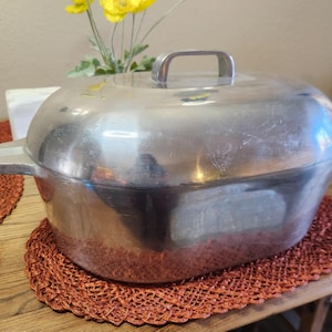 May include: A large, oval-shaped, aluminium roasting pan with a lid and handle. The pan sits on a woven, reddish-brown placemat. Yellow flowers are visible in the background. The pan is designed for cooking and baking.