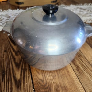 May include: A vintage aluminum pot with a lid and a black knob. The pot has two handles and a rounded shape. The pot is sitting on a wooden surface.