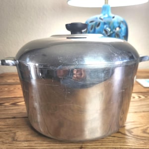 May include: A large, stainless steel cooking pot with a lid and two side handles. The pot has a black knob on top of the lid. The pot is sitting on a wooden surface.