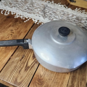 May include: A vintage aluminium saucepan with a lid and a black handle. The lid has a black knob. The saucepan is on a wooden surface.