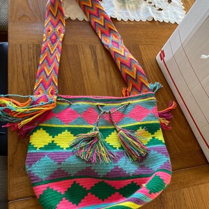 Op de afbeelding: Een kleurrijke, handgemaakte schoudertas met een levendig geometrisch patroon. De tas heeft een geweven ontwerp in roze, groen, geel en blauw, met bijpassende kwastjes en een patroonband. De tas is ongeveer 30 cm breed.