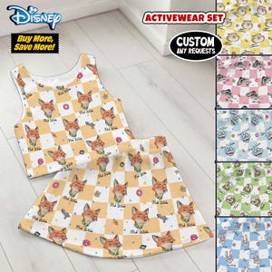 May include: A two-piece activewear set featuring a Nick Wilde design from Disney's Zootopia. The set includes a crop top and skirt with a repeating pattern of Nick Wilde's face, donuts, and guns on a yellow and white checkered background. The set is labeled "ACTIVEWEAR SET".