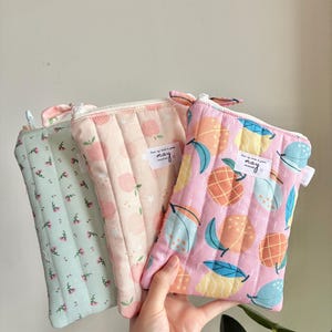 Pode incluir: Três bolsas acolchoadas com zíper em cores pastel. Uma é verde claro com um padrão floral, outra é rosa com desenhos de pêssegos e a terceira é rosa com ilustrações de frutas. Cada bolsa tem uma pequena etiqueta que diz "may."