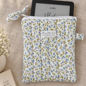 May include: A quilted fabric sleeve for an e-reader featuring a white background with a pattern of small blue flowers and green leaves. The sleeve has a white zip with a matching fabric pull and a label reading May Studio from my hands to yours.