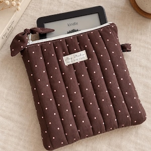 May include: A brown quilted fabric sleeve with small pink polka dots, featuring a white zip closure with a matching fabric bow pull. A rectangular label reads "Mag Studio from my hands to yours". A black Kindle e-reader is tucked inside.