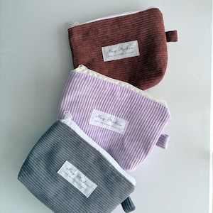 May include: Three corduroy pouches in burgundy, lavender, and grey, each with a white zip and a small fabric loop. Each pouch has a white label with the text "May Studio" and "from my hands to yours."