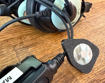 May include: Black aviation headset with a black control box and a small, black, square-shaped accessory. The headset has light green ear cushions and is connected to the control box and accessory by black cords. The accessory has a silver Apple logo.