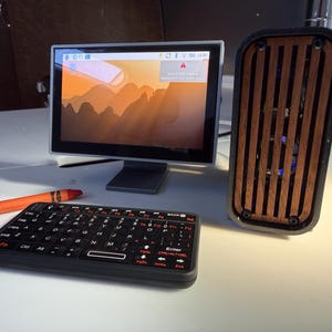 May include: A small computer setup featuring a monitor displaying a landscape image, a black keyboard, and a speaker with a wooden front. An orange crayon is also present. The monitor is approximately 6 inches wide.