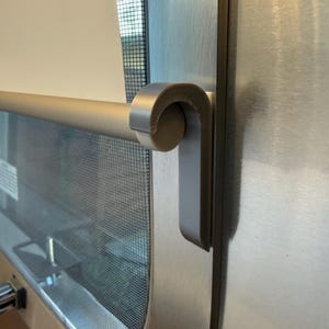 May include: Close-up of a brushed metal door with a curved handle. The handle is a light, neutral color and has a rounded end. A screen is visible behind the handle, and the door frame has a metallic finish.