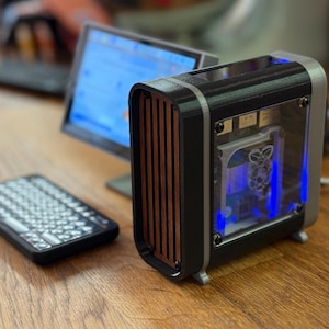 May include: A small, custom-built computer case with a black and silver frame, wood-grain front panel, and clear side panel. A small keyboard and monitor are also visible on a wooden desk. The case has blue internal lighting.
